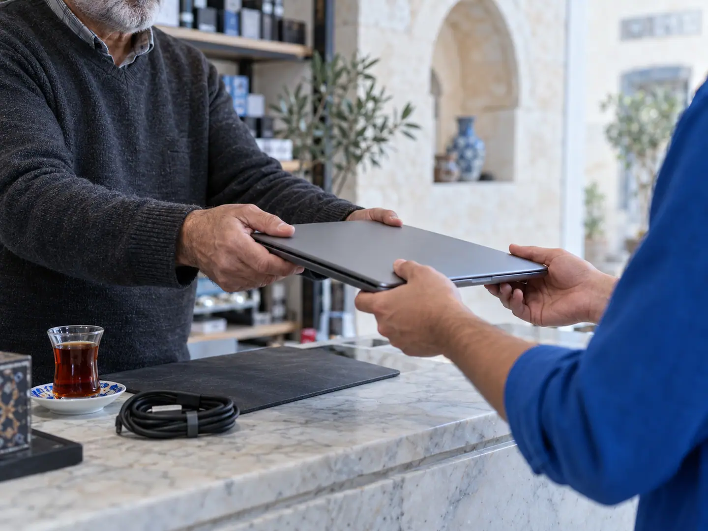 يدا بائع خلف منضدة رخامية تسلّمان لابتوبًا لزبون، وكأس شاي على المنضدة